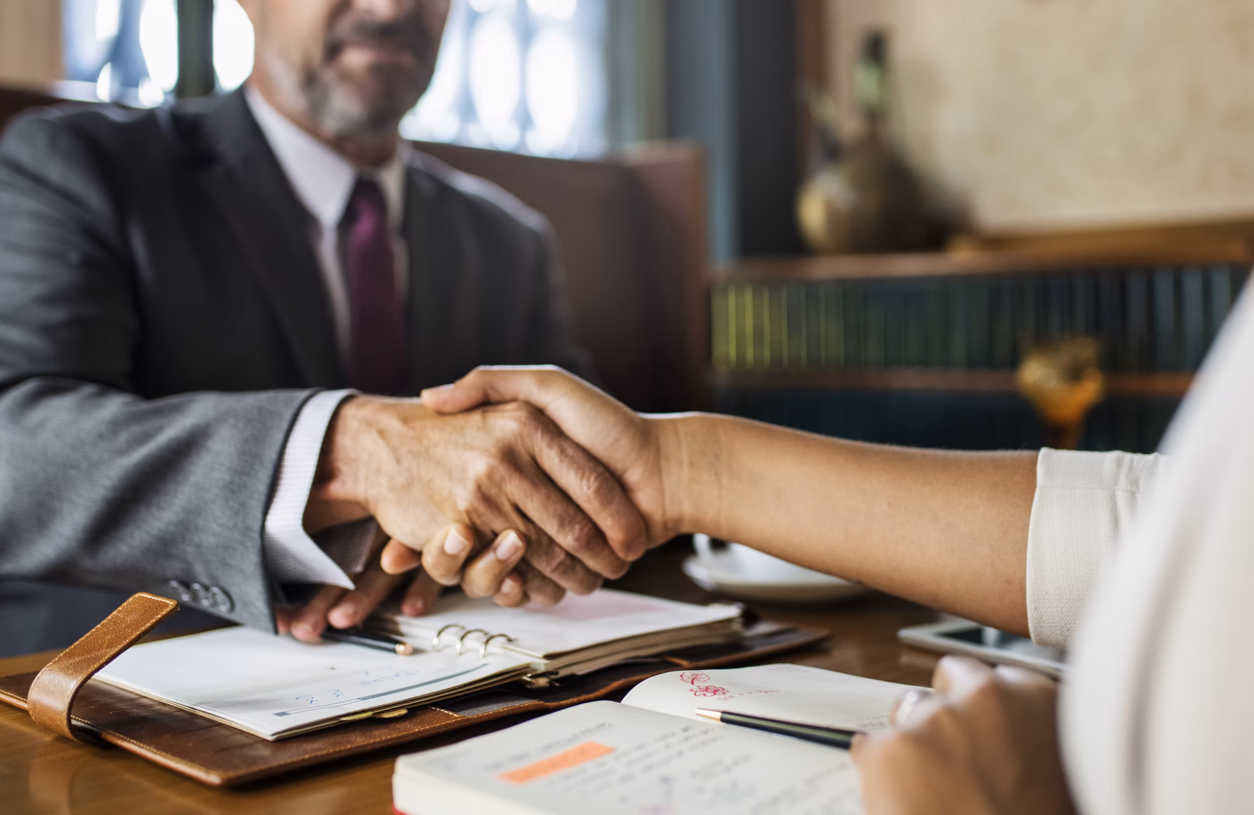 Two people shaking hands over a desk with open notebooks and planners.