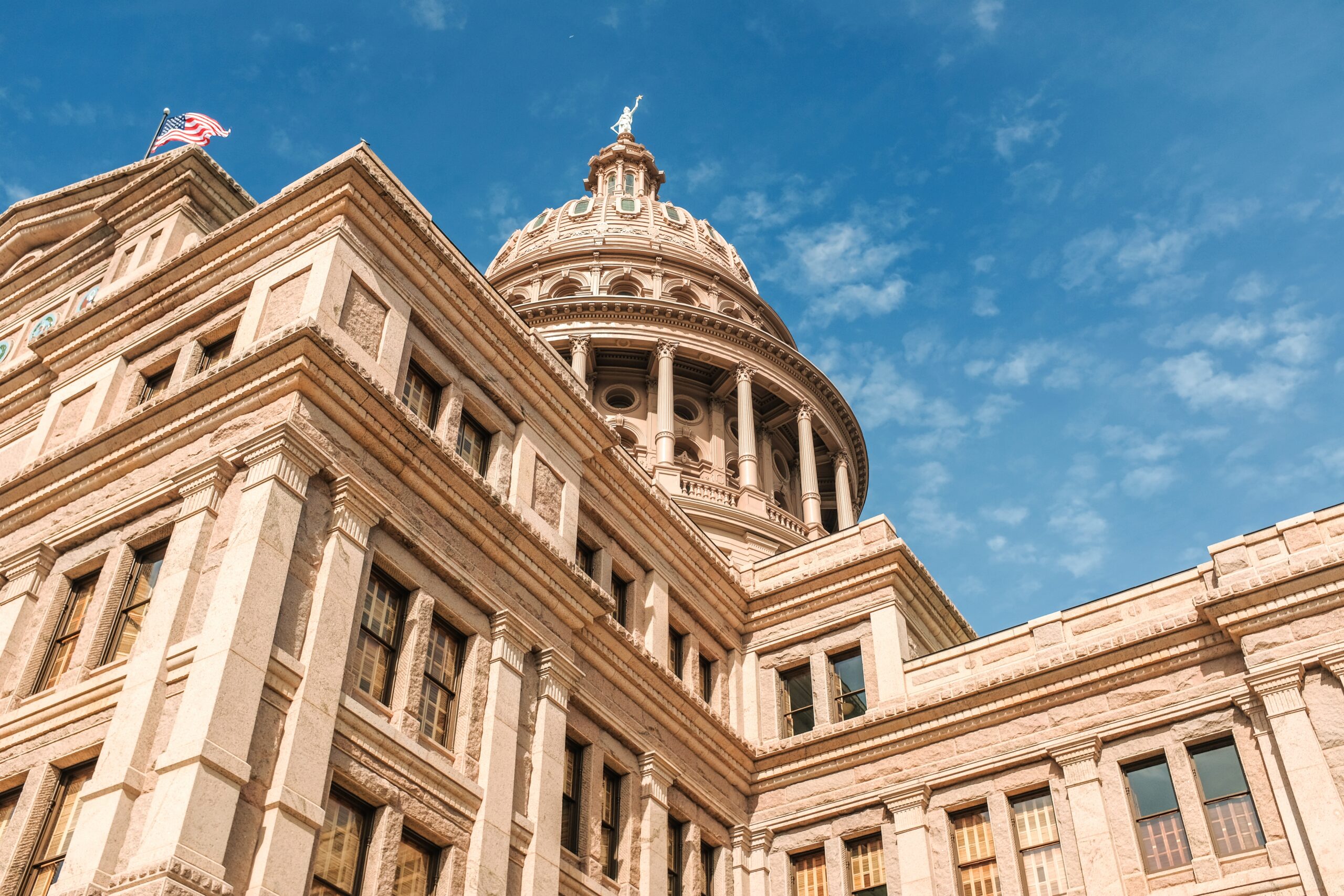 Granite capitol building with dome and American flag against a bright blue sky
