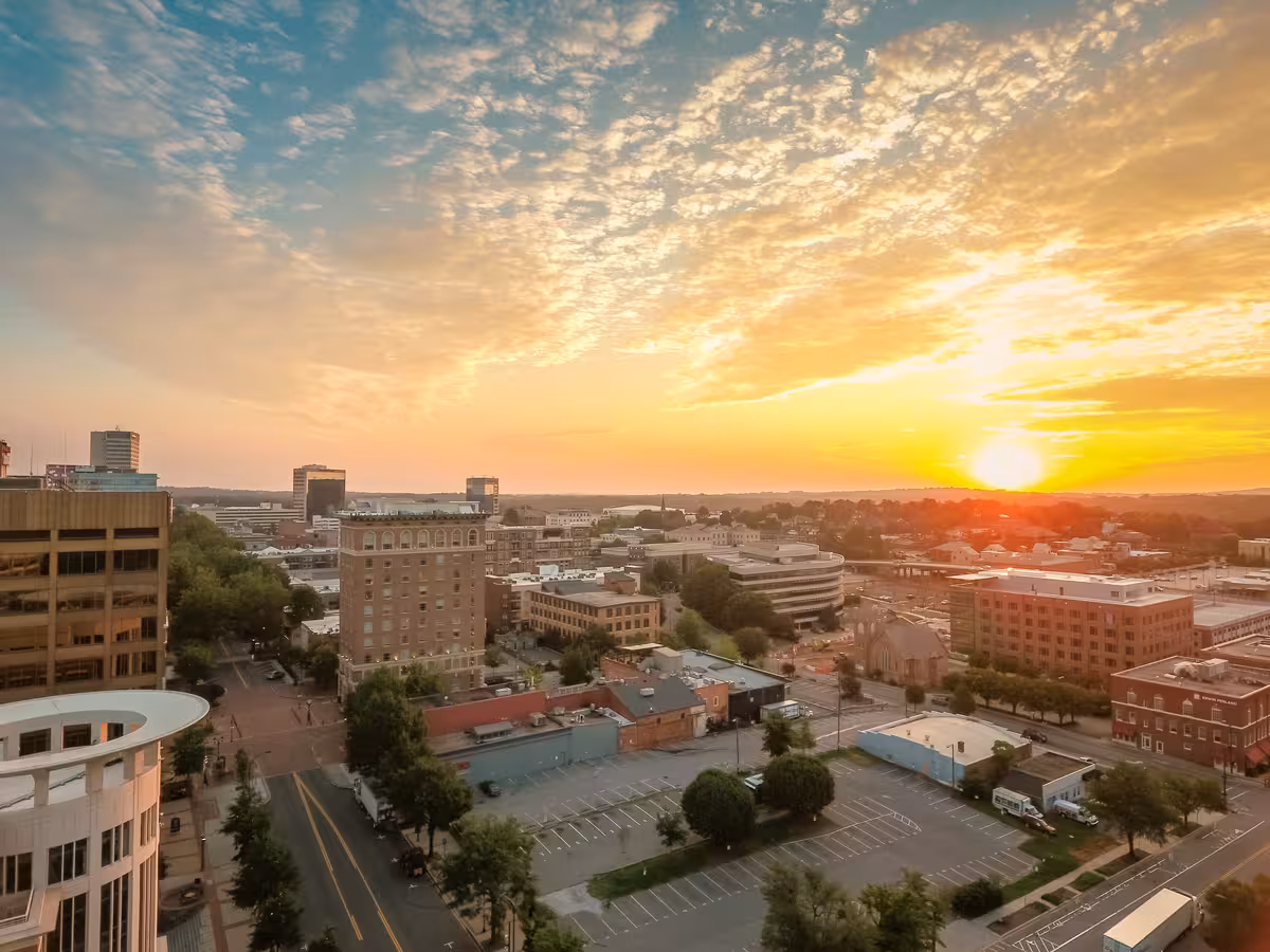 Aerial view of a downtown at sunset, with buildings, streets, and parking lots under a golden sky.