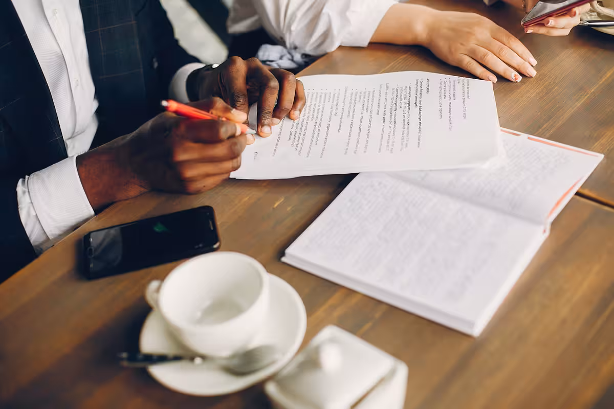 Person with a red pen reviewing documents at a table with an open notebook and coffee cup.