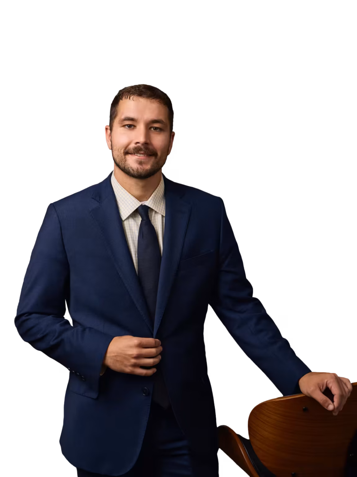 Smiling man in a navy suit and tie stands holding the back of a wooden chair.